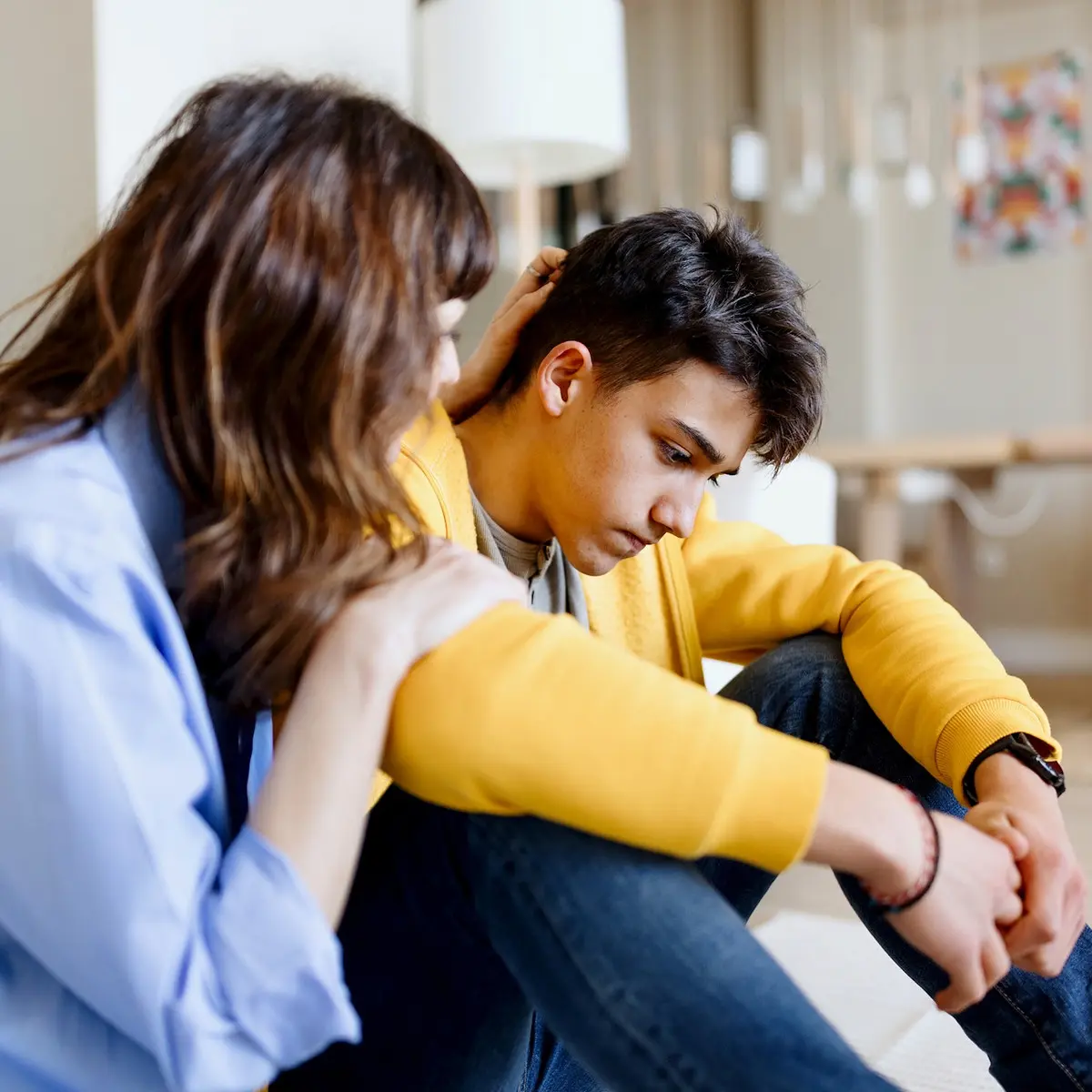 A mother comforts her depressed teen son as they sit on the living room floor